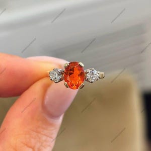 May include: A three-stone ring featuring a central oval-cut vibrant orange gemstone held in a four-prong setting, flanked by two round-cut clear white diamonds on each side, all mounted on a polished yellow gold band.