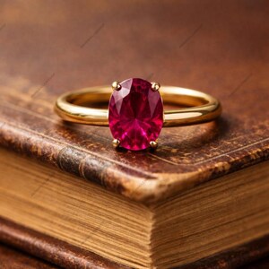 Può includere: Un anello in oro con una pietra preziosa rossa vibrante dal taglio ovale, fissata da quattro griffe. La fascia è liscia e lucida, caratterizzata da un design classico con una finitura brillante che riflette la luce sulla superficie sfaccettata.