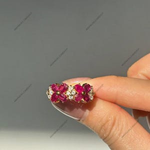 Può includere: Un anello in oro con due motivi a forma di fiore, ciascuno composto da quattro rubini rosa vivaci taglio ovale attorno a un punto centrale dorato, con piccoli diamanti bianchi rotondi incastonati tra i motivi floreali sulla fascia.