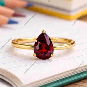 Pode incluir: Um anel de ouro com uma única pedra preciosa granada vermelho-escura em forma de pera, montada numa cravação de garras. A banda é lisa e polida, destacando o design facetado da pedra. O texto AMANAHGLEAMSHOP é visível na imagem.