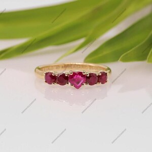 May include: A gold ring featuring five red gemstones set in a row. The center stone is heart-shaped, while the four flanking stones are round. The band is polished gold, and the metal surface shows faint engraved text along the inner curve.