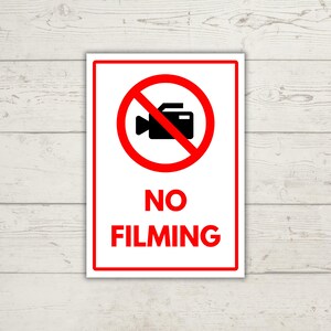May include: A rectangular sign with a white background and a red border. The sign features a black camera icon inside a red circle with a diagonal line through it, and the words "NO FILMING" in red.