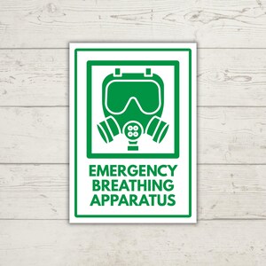 May include: A rectangular white sign with a green border featuring a green icon of a full-face respirator mask with two side filters. Below the icon, the text reads EMERGENCY BREATHING APPARATUS in bold green capital letters.