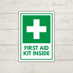Puede incluir: Un letrero rectangular con fondo blanco y borde verde. La sección superior presenta una gran cruz blanca sobre un cuadrado verde. La sección inferior contiene el texto FIRST AID KIT INSIDE en letras mayúsculas verdes en negrita.