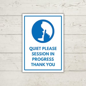 Könnte beinhalten: Rechteckiges Schild mit weißem Hintergrund und blauem Rand. Das Schild zeigt einen blauen Kreis mit der Silhouette einer Person, die zum Schweigen auffordert. Darunter steht der Text "QUIET PLEASE SESSION IN PROGRESS THANK YOU."