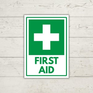 Puede incluir: Un letrero rectangular de primeros auxilios que presenta una cruz blanca centrada sobre un fondo verde. Debajo de la cruz, el texto FIRST AID está impreso en letras mayúsculas verdes en negrita. El letrero tiene un borde verde fino.