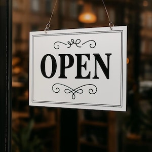 Könnte beinhalten: Ein rechteckiges weißes Schild mit dem Wort "OPEN" in fetten schwarzen Buchstaben. Das Schild hat einen schwarzen Rahmen und dekorative Schnörkel oben und unten. Es hängt an dünnen Drähten.