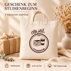 Könnte beinhalten: Eine beigefarbene Stofftasche aus Baumwolle mit einem dunkelbraunen, kreisförmigen Aufdruck, der einen Bücherstapel, einen Kaffeebecher, das Jahr 2026 sowie den Text "Ab jetzt" und "STUDENTIN" zeigt. Die Tasche hat zwei Tragegriffe.