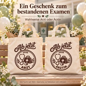 Könnte beinhalten: Zwei cremefarbene Stofftaschen mit einem dunkelbraunen kreisförmigen Aufdruck, der ein Stethoskop, eine Arzttasche, das Jahr 2026 sowie den Text 'Ab jetzt ARZT' auf der einen und 'Ab jetzt ÄRZTIN' auf der anderen Tasche zeigt, gefüllt mit Blumen.