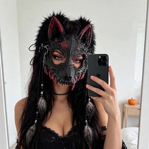 Peut inclure: Un masque de loup noir et rouge avec des oreilles pointues, des dents acérées et des détails texturés complexes. Le masque est orné de longue fausse fourrure noire, de chaînes pendantes et de plumes grises. Il couvre le haut du visage.