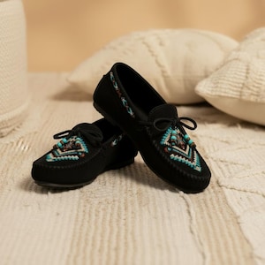 May include: A pair of black suede moccasin-style shoes with intricate turquoise, brown, and white geometric embroidery on the toe and sides. The shoes have a tie closure and are displayed on a textured surface.