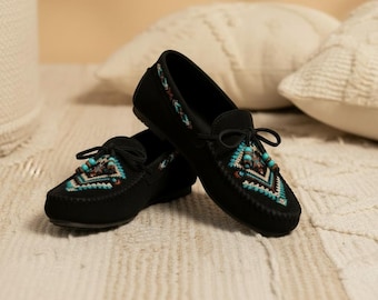 Può includere: Un paio di scarpe mocassino in camoscio nero con intricati ricami geometrici turchese, marrone e bianco sulla punta e sui lati. Le scarpe hanno una chiusura con lacci e sono esposte su una superficie strutturata.