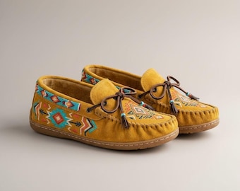 Può includere: Un paio di scarpe mocassino giallo senape con intricati ricami e perline di ispirazione nativa americana. Le scarpe presentano lacci in pelle marrone, nappe e una suola cucita. Il design include motivi geometrici in turchese, rosso e arancione.