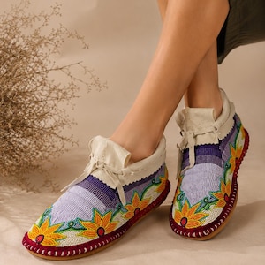 May include: Beaded moccasin-style shoes with a cream-colored leather collar and laces. The shoes feature a floral design with yellow, orange, and green flowers on a light purple background. The soles are red.