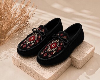 Può includere: Un paio di mocassini in camoscio nero con un pannello decorativo sulla parte superiore caratterizzato da un motivo geometrico in filo rosso, bianco e nero. Ogni scarpa include un laccetto nero sul davanti e una cucitura sulla punta arrotondata.