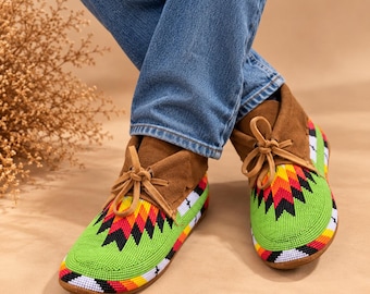 Può includere: Mocassini in camoscio marrone con un vivace motivo di perline sulla punta e sui lati. La lavorazione a perline presenta un design geometrico in verde brillante, giallo, arancione, rosso, nero e bianco. Ogni scarpa ha lacci in pelle.