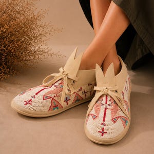 May include: A pair of cream-colored suede moccasins featuring intricate beadwork in shades of red, turquoise, and white. The shoes have a lace-up front closure with a distinctive pointed tongue design and a flat sole, showcasing detailed geometric patterns.