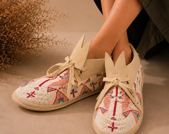 Può includere: Un paio di mocassini in camoscio color crema con intricate perline nei toni del rosso, turchese e bianco. Le scarpe presentano una chiusura frontale con lacci, un design a linguetta appuntita e una suola piatta con dettagliati motivi geometrici.