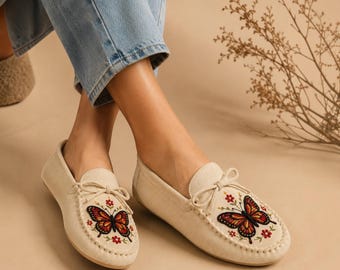 Può includere: Un paio di mocassini color crema con un fiocco decorativo sulla tomaia e un intricato ricamo di una farfalla monarca circondata da piccoli fiori rossi. Le scarpe presentano una finitura morbida e testurizzata con punta arrotondata.