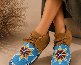 Può includere: Un paio di mocassini in camoscio marrone con una tomaia in perline blu brillante caratterizzata da un motivo a stella multicolore in rosso, giallo e bianco. Le scarpe presentano lacci in camoscio abbinati e un bordo di perline bianco e nero.