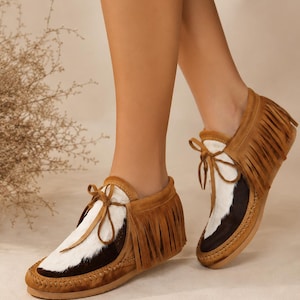 May include: Brown suede moccasin shoes with fringe detailing. The shoes feature a cowhide design with white and black patches. The shoes have a leather tie and a stitched sole. These shoes are a stylish addition to any wardrobe.