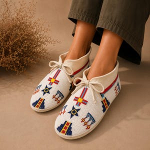May include: A pair of off-white ankle boots featuring intricate beadwork in red, blue, and yellow geometric patterns, including star and cross motifs. The boots have a lace-up front closure with matching off-white laces and a rounded toe design.