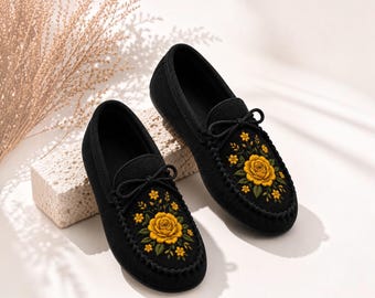 Può includere: Un paio di mocassini neri senza lacci con un fiocco decorativo sulla tomaia e un intricato ricamo di una grande rosa gialla circondata da fiori gialli più piccoli e foglie verdi sulla parte superiore di ogni scarpa.