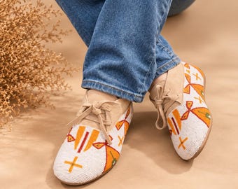 Può includere: Scarpe stringate in camoscio beige con intricati motivi di perline bianche, arancioni e gialle sulla punta e sui lati. La lavorazione a perline include forme geometriche e motivi a croce. Le scarpe hanno lacci beige abbinati.