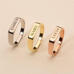 Può includere: Tre anelli in metallo con parte superiore rettangolare piatta, ognuno con un'incisione personalizzata. Il primo anello è color argento con il nome EMBER, il secondo è color oro con il nome MABEL e il terzo è color oro rosa con il nome LUCIA.