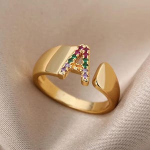 Op de afbeelding: Een goudkleurige open ring met een hoofdletter A als middelpunt. De letter A is versierd met kleine, veelkleurige edelstenen in tinten rood, groen, geel en paars, wat zorgt voor een levendig, regenboogachtig effect op het oppervlak.