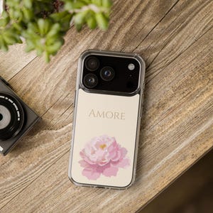 Op de afbeelding: Transparante telefoonhoes met een crèmekleurige inzet met het woord "AMORE" en een roze pioenroosontwerp. De hoes ligt op een houten oppervlak, naast een camera en een plant.