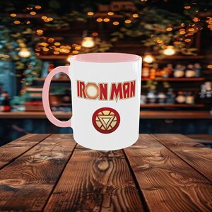 May include: White ceramic mug with a pink interior and handle. The mug features the words "IRON MAN" in a bold, red and gold font, along with the Iron Man logo. The mug is set on a wooden table.