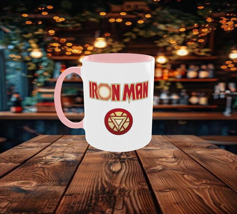 May include: White ceramic mug with a pink interior and handle. The mug features the words "IRON MAN" in a bold, red and gold font, along with the Iron Man logo. The mug is set on a wooden table.