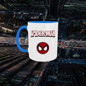 May include: White ceramic mug with a blue handle and interior. The mug features the words "SPIDER-MAN" in red and white, with a red and black spider symbol below the text. The mug is set against a cityscape background.