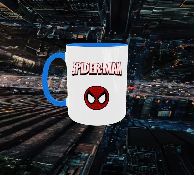 May include: White ceramic mug with a blue handle and interior. The mug features the words "SPIDER-MAN" in red and white, with a red and black spider symbol below the text. The mug is set against a cityscape background.