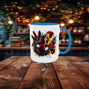 May include: White ceramic mug with a blue interior and handle, featuring a colorful graphic of Iron Man. The design shows Iron Man's hand and helmet with red, gold, and blue accents. The mug is set on a wooden table.