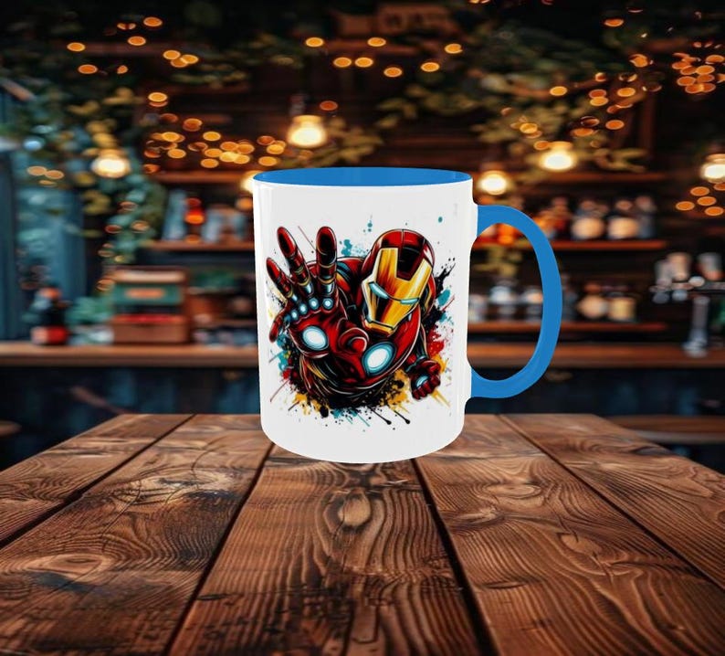May include: White ceramic mug with a blue interior and handle, featuring a colorful graphic of Iron Man. The design shows Iron Man's hand and helmet with red, gold, and blue accents. The mug is set on a wooden table.