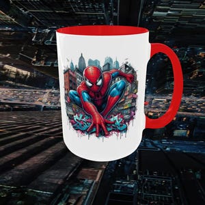 May include: White ceramic mug with a red interior and handle. The mug features a colorful illustration of Spider-Man in a city setting. The design includes graffiti-style lettering at the bottom of the image.