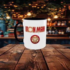 May include: A white ceramic mug with a black handle and interior. The mug features the words "IRON MAN" in red and gold lettering, along with a red and gold circular emblem. The mug is set against a blurred background.