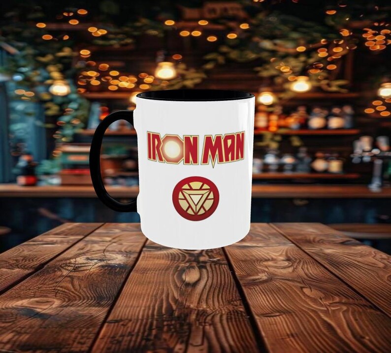 May include: A white ceramic mug with a black handle and interior. The mug features the words "IRON MAN" in red and gold lettering, along with a red and gold circular emblem. The mug is set against a blurred background.