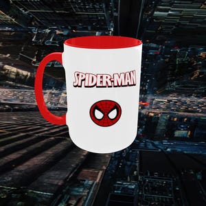 May include: A white ceramic mug with a red interior and handle. The mug features the text "SPIDER-MAN" in red and a red and black spider symbol. The mug is set against a blurred cityscape background.
