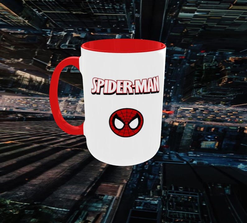 May include: A white ceramic mug with a red interior and handle. The mug features the text "SPIDER-MAN" in red and a red and black spider symbol. The mug is set against a blurred cityscape background.