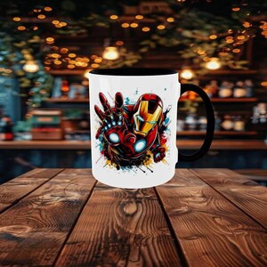 May include: White ceramic mug with a black handle and interior. The mug features a colorful graphic of Iron Man's hand and helmet. The design includes red, gold, and blue accents, with paint splatter effects. The mug is on a wooden table.