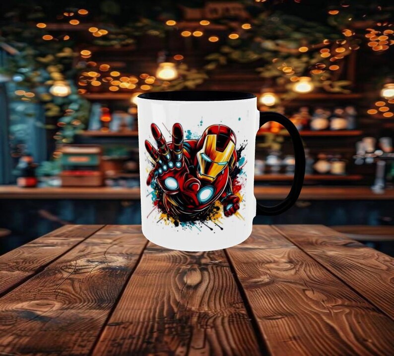 May include: White ceramic mug with a black handle and interior. The mug features a colorful graphic of Iron Man's hand and helmet. The design includes red, gold, and blue accents, with paint splatter effects. The mug is on a wooden table.