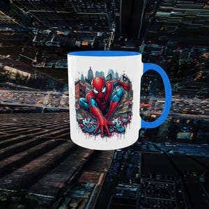 May include: White ceramic mug with a blue interior and handle. The mug features a colorful illustration of Spider-Man in a crouching pose, set against a cityscape background. The design includes graffiti art elements.
