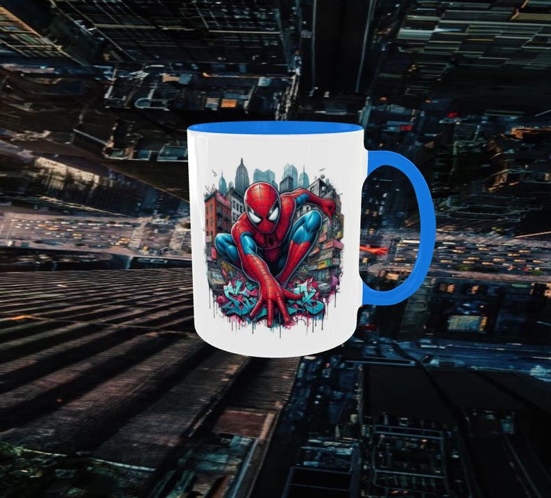 May include: White ceramic mug with a blue interior and handle. The mug features a colorful illustration of Spider-Man in a crouching pose, set against a cityscape background. The design includes graffiti art elements.