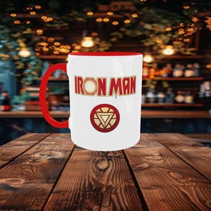 May include: White ceramic mug with a red interior and handle. The mug features the words "IRON MAN" in red and gold lettering, along with a circular emblem. The mug is sitting on a wooden table.