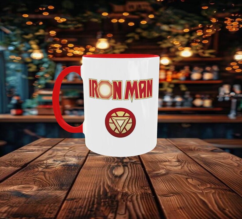May include: White ceramic mug with a red interior and handle. The mug features the words "IRON MAN" in red and gold lettering, along with a circular emblem. The mug is sitting on a wooden table.