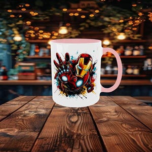 May include: A white ceramic mug with a pink interior and handle. The mug features a colorful graphic of Iron Man's hand and helmet. The design includes red, gold, and blue accents, with paint splatter effects. The mug is set on a wooden table.