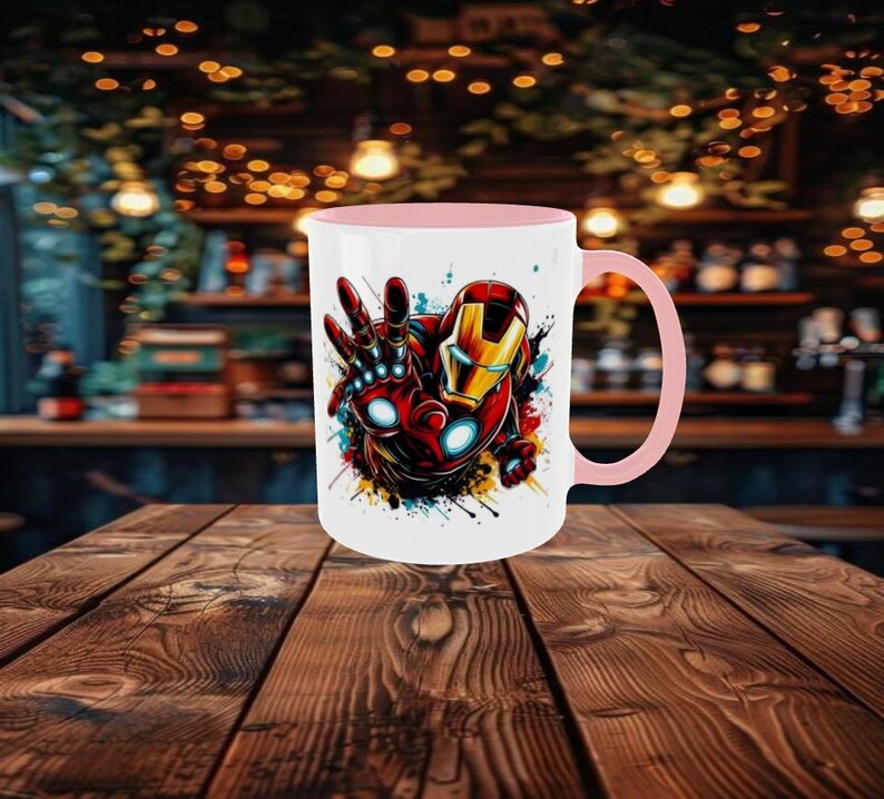 May include: A white ceramic mug with a pink interior and handle. The mug features a colorful graphic of Iron Man's hand and helmet. The design includes red, gold, and blue accents, with paint splatter effects. The mug is set on a wooden table.